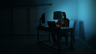 A hacker sitting at a computer screen wearing a mask with their hood up